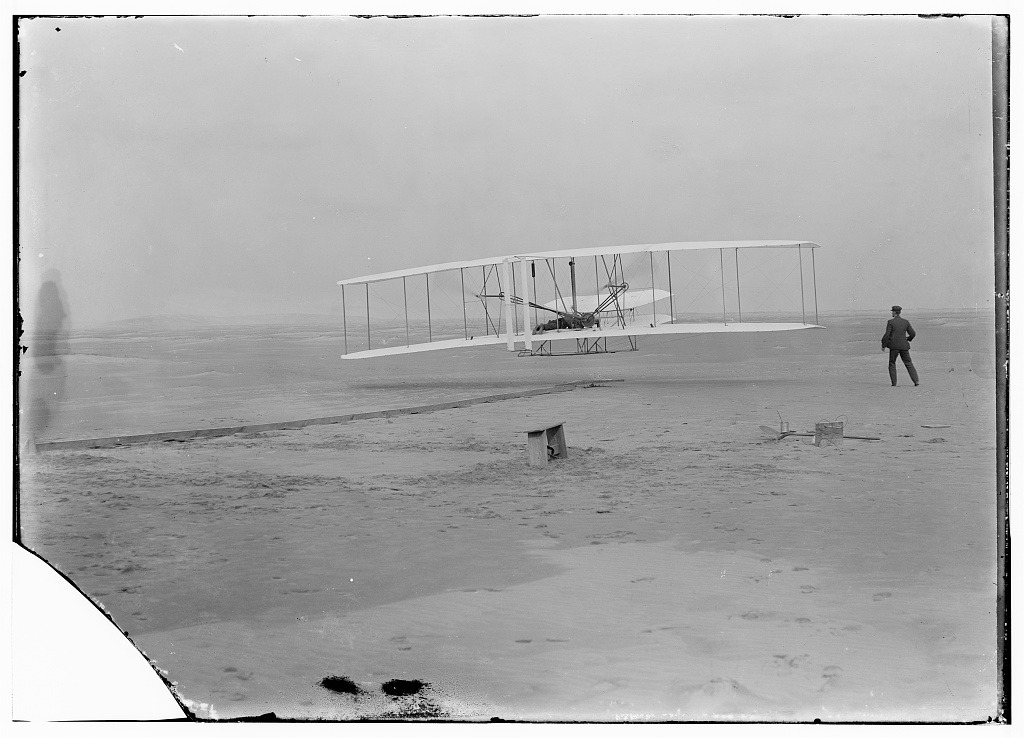 Orville Wright pilots the first powered flight as Wilbur runs alongside the airplane, 1903